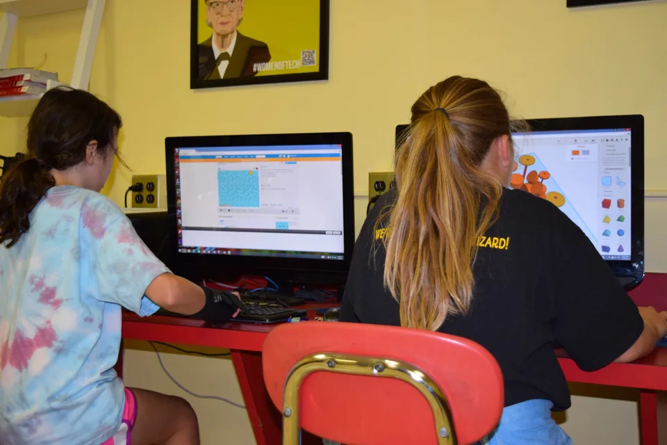 Two campers work at computers, one coding a game and one designing a 3D model