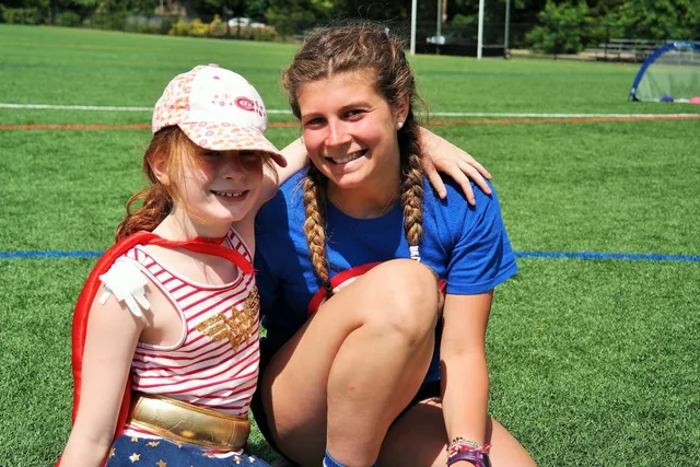 A counselor kneels on the field with her arm around a camper dressed as a superhero