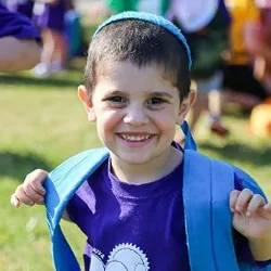 A smiling camper in a kippah holds the straps of his backpack