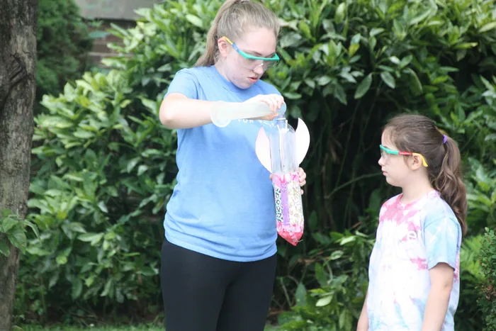 A counselor in safety goggles pours water into a camper's decorated bottle rocket